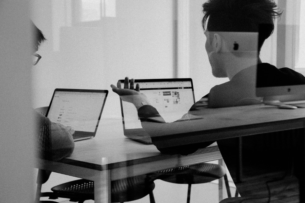 Black and white photo of two people collaborating at laptops through a glass office partition, one gesturing as if explaining or coaching